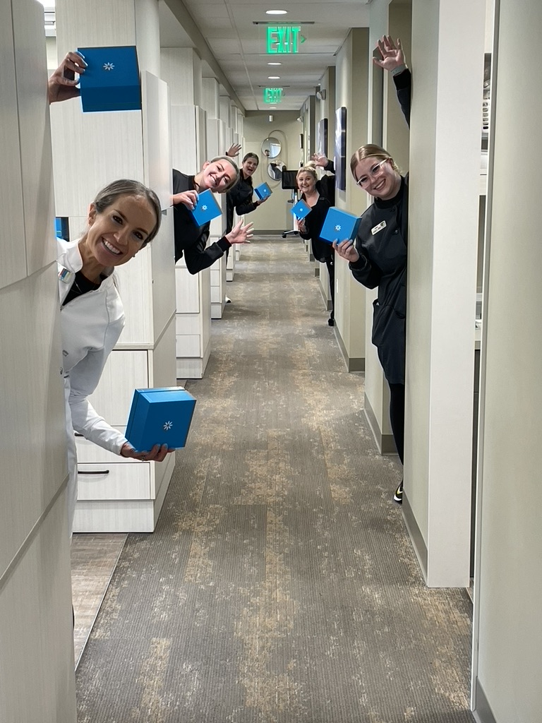 Smiling dental team playfully peeking out from exam rooms, holding blue Invisalign boxes in a clinic hallway.