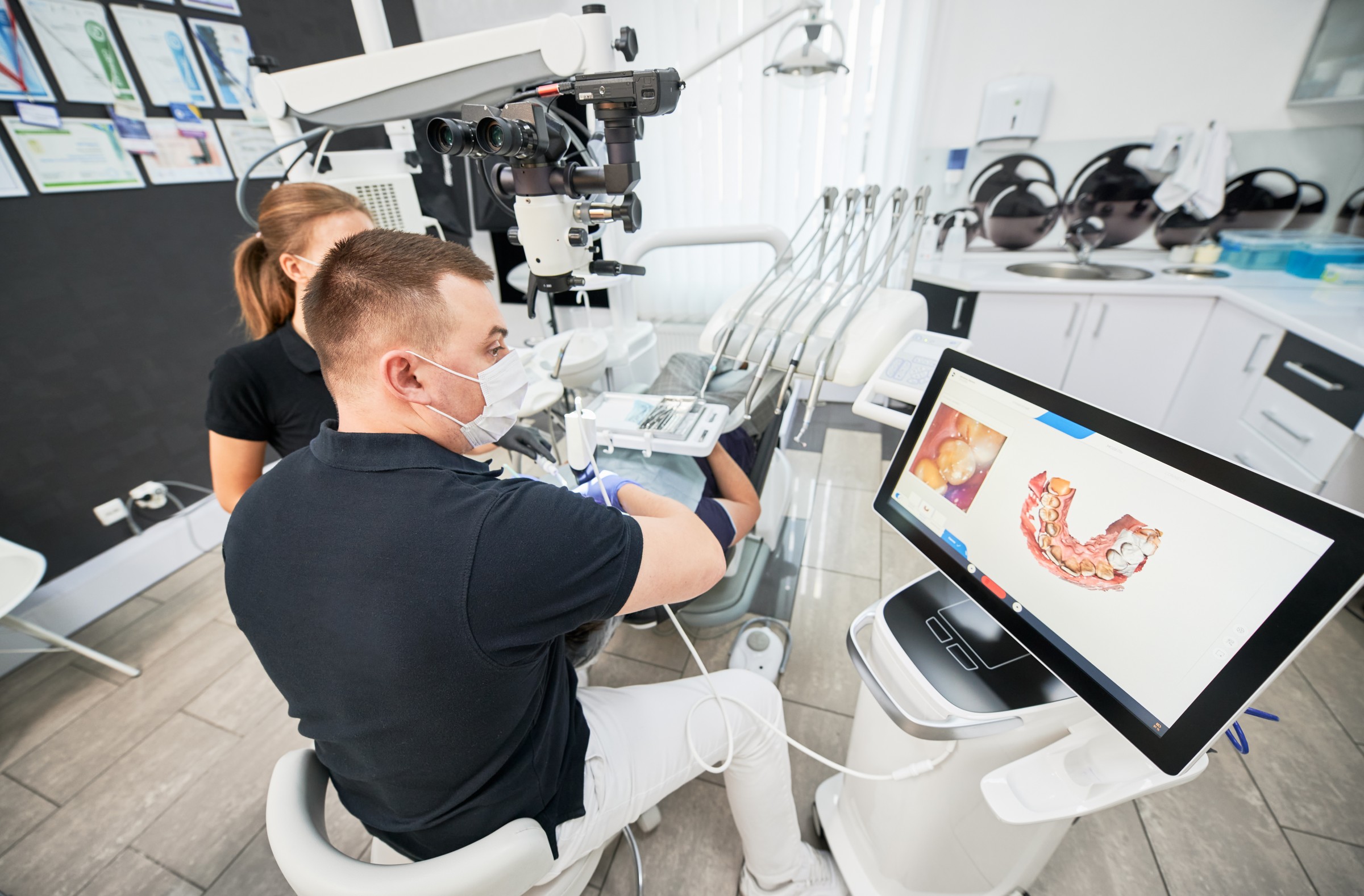 A dentist using a digital scanner on a patient, with a 3D dental image displayed on a monitor in a modern clinic.