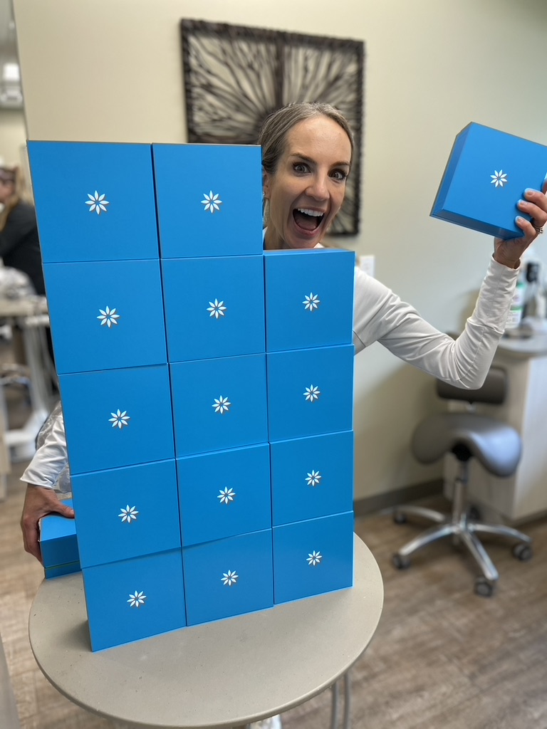 Excited woman in a dental office holding a stack of blue Invisalign boxes, smiling with enthusiasm.