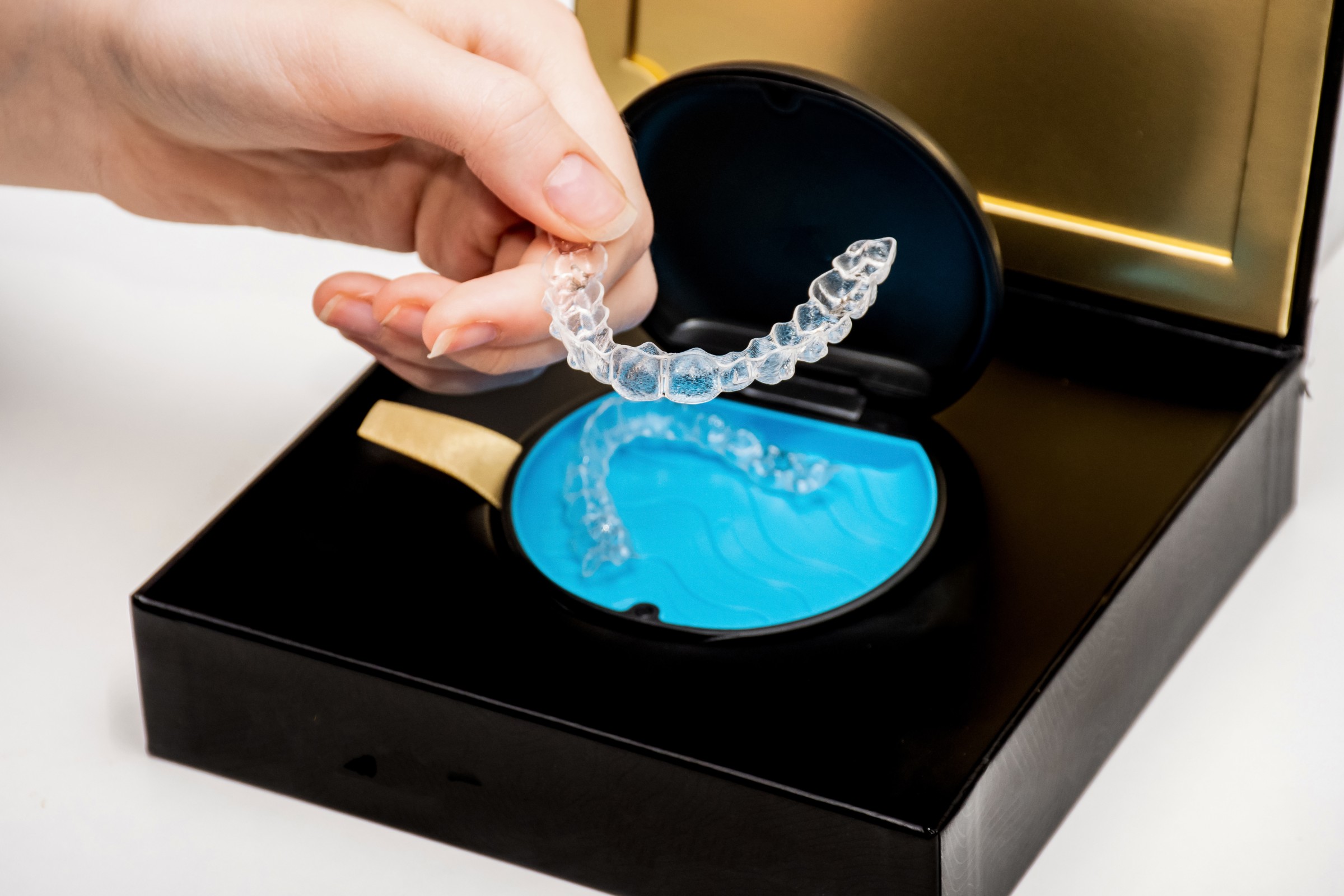 Hand holding a clear dental aligner above an open black and blue case inside a luxury box.